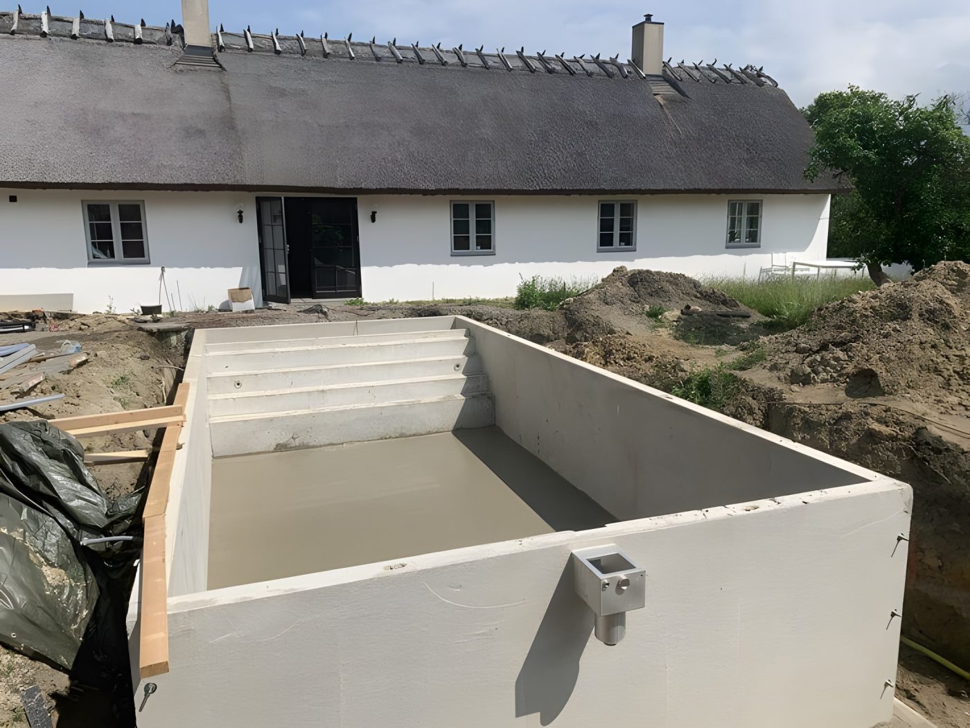 Betongpool under byggnation framför ett vitt hus med vasstak