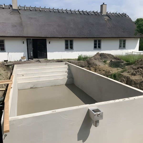Betongpool under byggnation framför ett vitt hus med vasstak